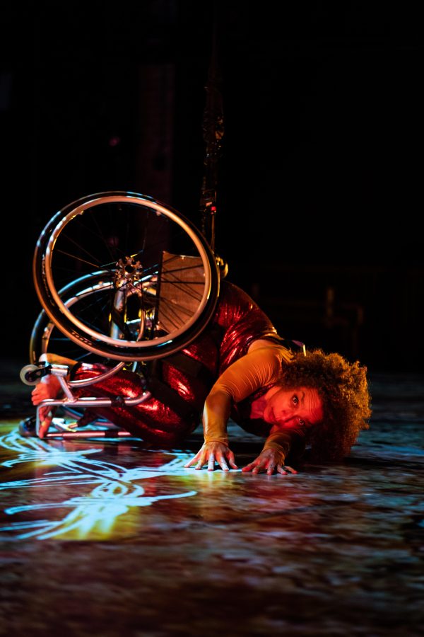 Alice Sheppard, a multiracial Black woman with coffee-colored skin and short curly hair, crouches onstage, legs and wheels curled toward her core, arms reaching toward the camera. Dappled purple and blue lighting covers Alice and the floor, as a projection of white and yellow barbed wire extends from under her. She peers intently beyond the camera, eyebrows raised.