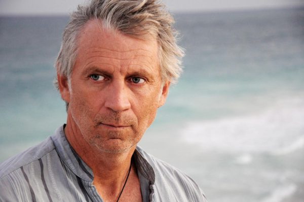 Close up portrait of a grey-haired man looking off to our left against a blurry ocean background.