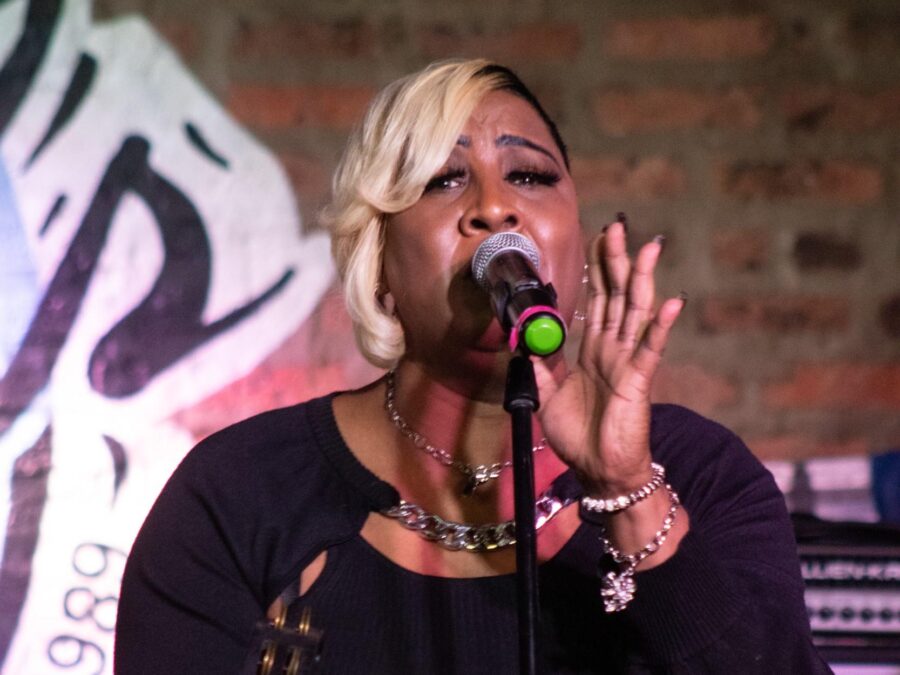 A Black woman performs into a microphone while holding a tambourine in her right hand.