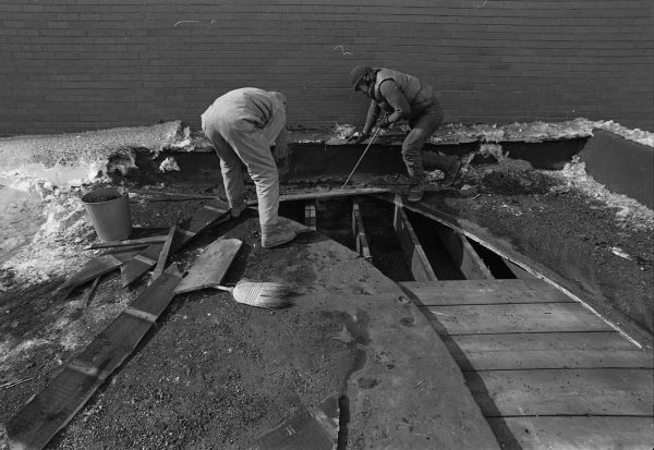 Two people in coveralls remove boards with crowbars, exposing the rafters below.