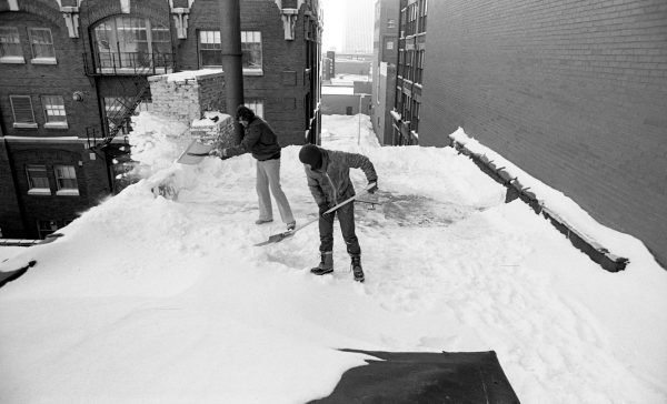 Two people shovel snow off the roof of a building.