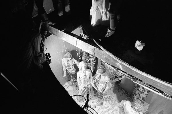 A group of people look up through a cut in a floor. The feet of several others viewing the group can be seen around the cut.
