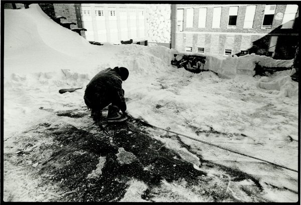 A person kneels on the roof of a building next to a large snow pile.