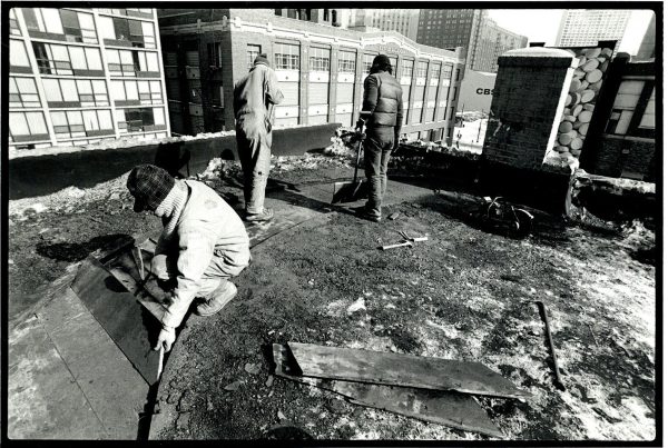 A crew of three people being to remove boards from underneath a top layer of the roof of a building.
