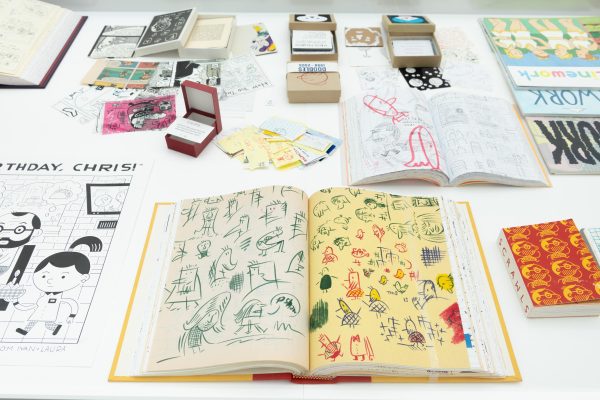 Books and other ephemera are displayed on a table. A few of the books are opened to different pages displaying a variety of sketches.