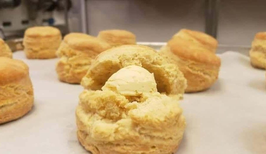 Ten baked biscuits scattered around a baking tray on parchment paper. The middle one closest to the foreground is broken open and has a large pat of butter in it.