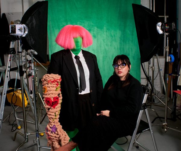 Ariella Granados, a woman with dark hair and glasses, sits in front of a green screen in a photography studio surrounded by props and equipment.