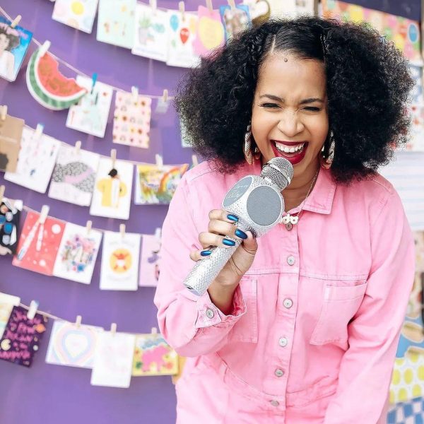 A Black woman dressed in pink holds a sparkly microphone. Her lips and eyes are partly open, suggesting she might be singing.