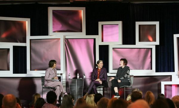 Three women sit on a stage surrounded by white-framed filled with pink, irridescent fabric