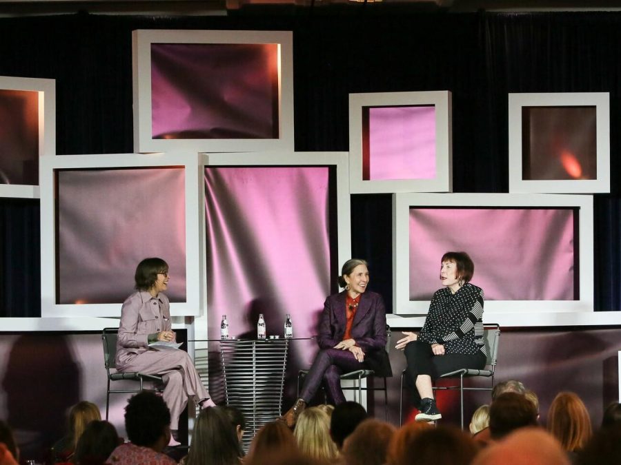 Three women sit on a stage surrounded by white-framed filled with pink, irridescent fabric