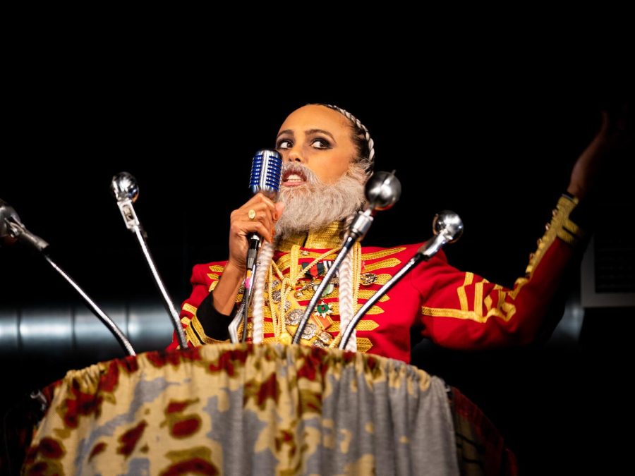 A female-presenting person with a fake grey beard and British red coat attire in front of a podium filled with microphones